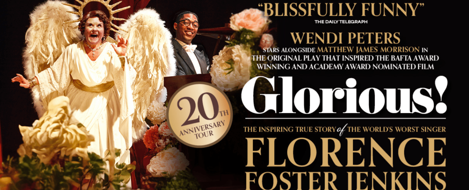 A woman with brown hair and a tiara wearing a pearl necklace and a pink top is singing in front of a pink background with an ornate golden frame, with a smiling man and woman next to her and one woman with her fingers in her ears