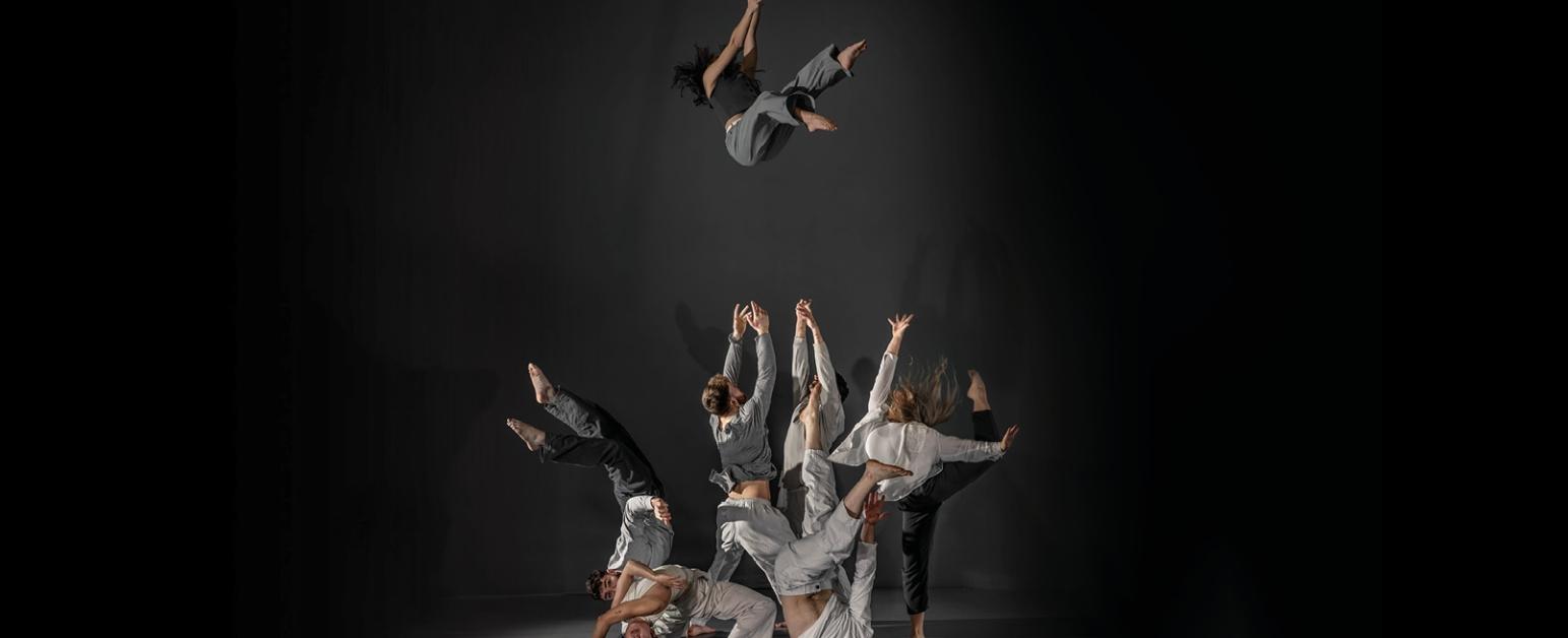 A performer falling into a group of performers with their arms raised behind other performers on the floor who are contorted in various positions, all wear black, grey and white