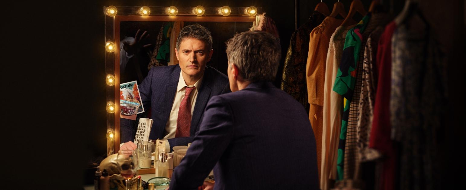 Tom Chambers looking in a mirror which is surrounded by lights. He is in a dressing room with costumes hanging beside him. The reflection shows an eerie hand reaching towards him. 