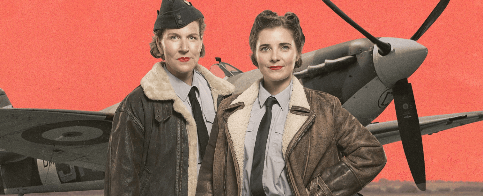 Two women stand in front of a spitfire wearing suits and early 20th century flying jackets