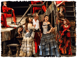 Multiple performers in dresses surround a man in a white top