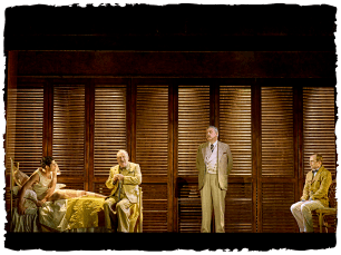 An actor on a bed, three other actors standing aorund wearing suits