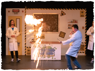 A illar of fire blazes between two performers dressed as scientists