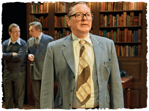 Man in suit and glasses looking serious, with two men stood whispering behind him