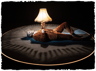 Man in orange suit lying centre stage with the glow of a lamp