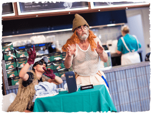 Actors in a cafe. One sitting behidn a table and one standing next to it in a ginger wig and hat