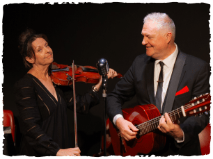 Man in grey suit playing guitar, smiling at woman in black dress whom is playing violin