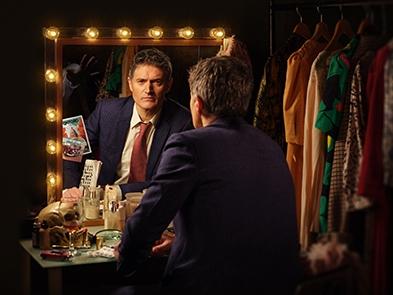 Tom Chambers looking in a mirror which is surrounded by lights. He is in a dressing room with costumes hanging beside him. The reflection shows an eerie hand reaching towards him. 
