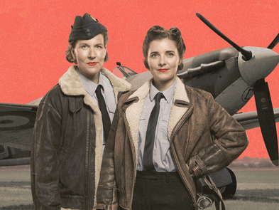 Two women stand in front of a spitfire wearing suits and early 20th century flying jackets