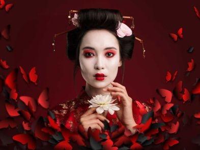 A woman with white and red makeup holds a white flower and stands amongst red petals