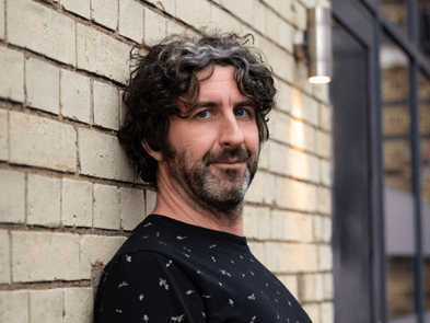 Mark Watson smiling while stood in front of a brick wall