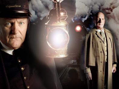 A signalman in a black uniform and hat holds a gaslight up with a man in the background wearing a trenchcoat. Both are looking straight ahead, with very serious expressions. There is a steam train and smoke.