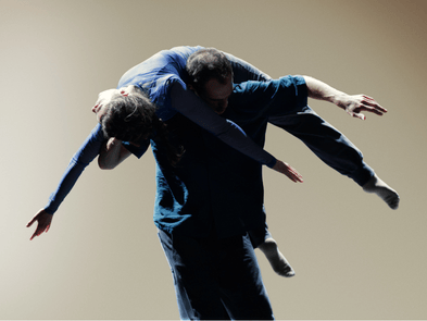 James Wilton Dance - two dancers against a beige background. Both are wearing blue and a male dancer is holding another dancer over his shoulder.