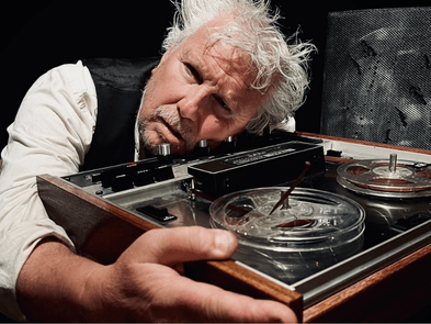 A man with scruffy grey hair and a beard looks worried as he rests his head on and clutches a tape recorder. 