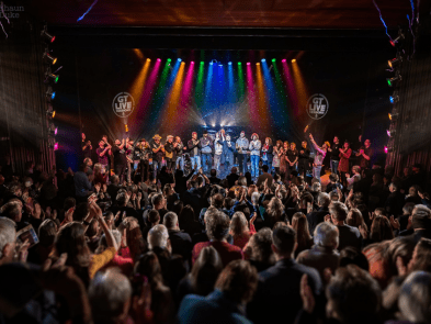 Lots op people stood on stage, audience giving a standing obviation. Bright colourful lighting.