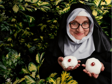 A nun in a habit hiding in the bushes holding two iced buns with cherries on top and smiling in a mischievous way.