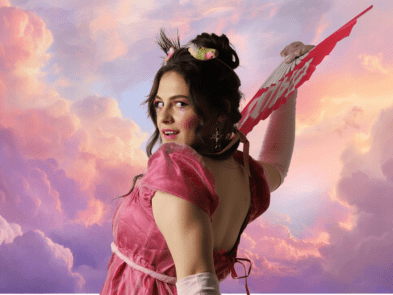 Woman in pink velvet regency style dress, hair up  and holding a fan. Her back is to the viewer but she has her head turned to face the viewer with a smile. The background is pink clouds/sky. 