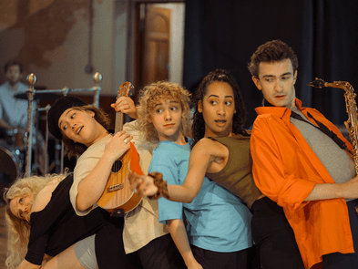 Five people stand in a row. One is holding a saxophone, one a ukulele, and they are leaning back on each other in a line