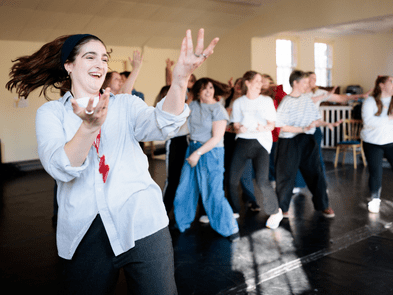 Woman with dark brown hair, white and blue stripe shirt and jeans is smiling whilst holding up her hands. There is a group of people behind her, dressed casually and all standing in different positions. 