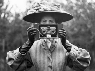 black and white image of a woman in Edwardinan clothing and a big hat, holding a camera. 